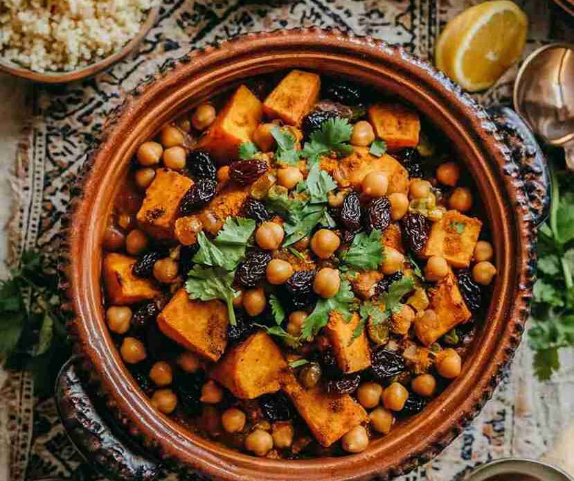 A close-up of a traditional Moroccan vegan tajine filled with sweet potatoes, chickpeas, and raisins, garnished with fresh cilantro. The dish is served in a rustic clay tajine pot placed on an intricately patterned tablecloth. Surrounding the tajine are complementary ingredients, including a bowl of fluffy couscous, turmeric powder, and lemon wedges, creating a warm, inviting presentation.